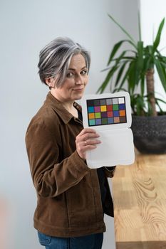 Pretty woman holds color checker or special target for color adjustment while standing in office interior. Gray haired woman showing color palette to adjust the color.