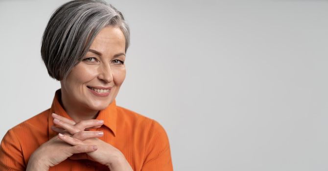 Happy aged woman smiling fingers crossed. Lady with bob hairstyle in orange blouse, head and shoulders portrait. Wisdom concept. Horizontal blank or template with copy spase at right side.