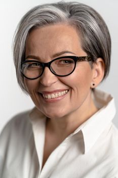 Pretty gray haired woman smiling at camera. Mature woman in eyeglasses and white shirt broadly smiles. Business concept. Education concept. Close up portrait.