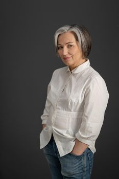 Gray-haired mature woman smiles holding hands in pockets while looking at camera. Charming caucasian woman in white shirt posing on a gray background.