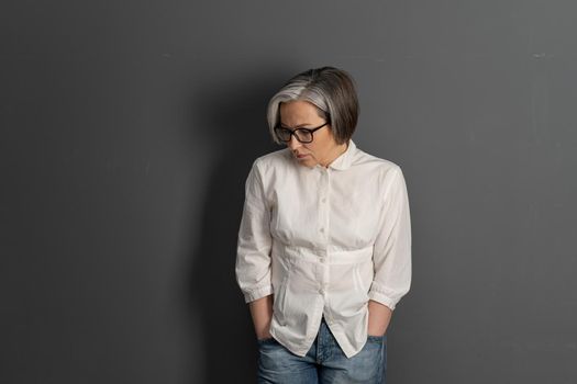 Sad middle-aged woman hunched looking down thoughtfully. Silver-haired stylish intelligent businesswoman with care hairstyle on grey background. Copyspace at both sides. Lonely concept.