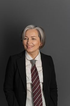 Smiling middle aged businesswoman on a gray background. Mature gray-haired caucasian woman wearing black jacket with tie looks at side smiling broadly. Business concept.