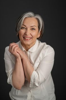 Joyful Caucasian woman smiles broadly while looking at camera and clasped her hands. Enthusiastic gray-haired mature woman in a white shirt posing in studio.