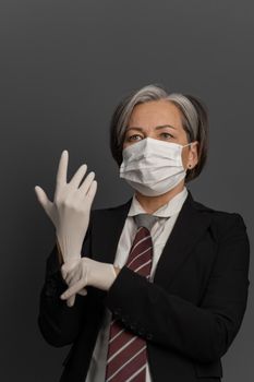 Businesswoman in medical mask putting on white disposable gloves. Wise mature woman complies with safety measures due to virus outbreak.