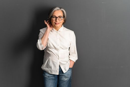 Pretty mature woman corrects medium-length hairstyle coquettishly. Adorable Gray-haired Caucasian model in jeans posing on gray background. Copy space or textspace at right side.