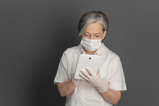 Gray-haired female doctor working with notepad. Caucasian woman wearing white protective uniform, mask and goggles using digital tablet. Isolated on gray back with copyspace on left.