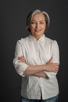 Charming middle-aged woman gently smiles standing crossed arms. Pretty lady looking at camera on gray background.