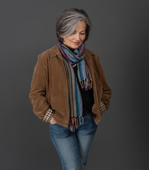 Creative gray-haired woman in casual on gray, portrait. Lady in jacket, colorfull scarf and jeans smiles looking down while standing hands in pockets.