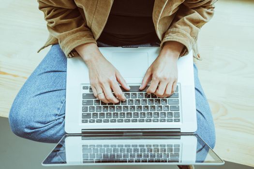 Woman types at laptop keyboard sitting on wooden windowsill. Woman in casual using computer. High angle view. Close up shot. Tinted image.