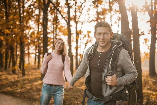 Two Travelers Walks Along Forest Trail Holding Hands, Selective Focus On Smiling Caucasian Man Looking At Camera While Standing In Back Sunlight, Guy And Girl With Backpacks Are Hiking Outdoors