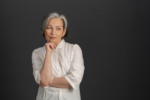 Thoughtful mature woman thinks touching by hand of her chin while looking at side. Gray-haired lady in white shirt isolated on gray background. Textspace at right side.