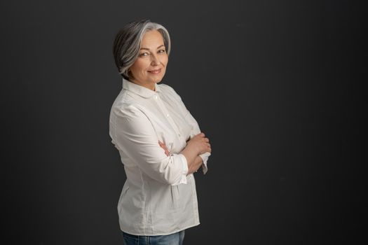 Pretty white-haired woman smiles looking at camera with arms crossed. Stylish caucasian businesswoman isolated on dark gray background. Textspace at right side. Business concept.