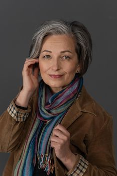 Stylish graying Woman charmingly smiles looking at camera. Caucasian mid aged beauty dressed in brown corduroy jacket with multi-colored scarf touches her face with hand. Anti-aging skin care concept.