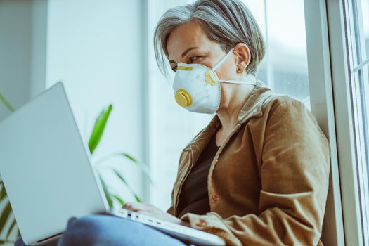 Mature woman in protective mask fp1 works laptop during quarantine. Grey haired businesswoman uses computer sitting on sill leaning back against the window indoor. Bottom side view. Tinted image.