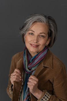 Charming mature woman in casual clothes smiles easily adjusting the collar of his jacket. Portrait of Caucasian lady in brown fustian jacket and colorful scarf coquettishly posing on gray background.