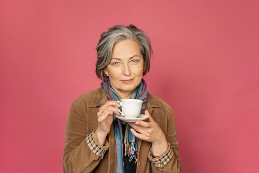 Charming Caucasian woman drinks coffee holding white cup. Pretty mature lady smiles slightly isolated on pink background. Beauty concept. Coffee time concept.
