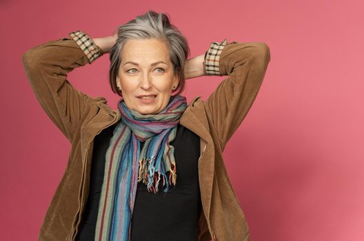 Graying mature woman making hairstyle correcting her hair by hands. creative woman gathered hair on the back of her head posing on a pink background.