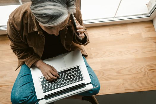 Grey haired woman speaking on mobile phone while working with laptop computer. Talking mature woman sitting on wide wooden window sill. Top view. Tinted image.