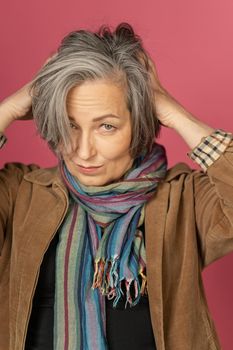 Graying mature woman making hairstyle, correcting her hair by hands. Creative lady gathered hair on back of her head posing in studio on pink background.