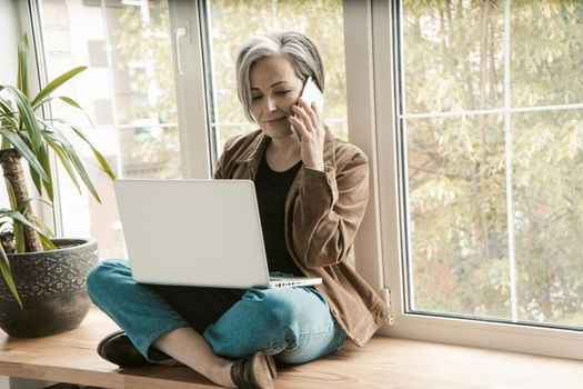 Charming woman talking on cell phone while working on laptop. Gray-haired aged woman sits cross-legged on wide windowsill, leaning her back against window. Tinted image.