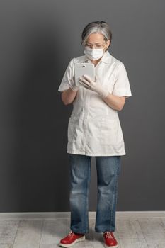 Doctor works with tablet computer. Serious mature woman in uniform and mask uses digital tablet standing full height in medical office.