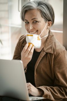 White-haired woman in age smiles taking off her mask and looking at the camera. Smiling woman with protective mask works laptop sitting on sill. Tinted image.