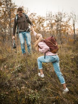 Backpackers Hiking On Hill, Intelligent Man Helps Woman Climb Hill, Man Stands On Top Holding Hiking Sticks In One Hand And Helping His Female Companion With Other Hand, Healthy Lifestyle Concept