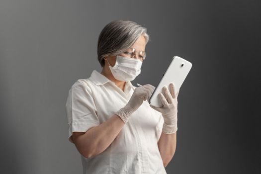 Side view of doctor using electronic tablet. Caucasian aged woman in protective mask works with digital gadget looking at screen on gray wall background. Focus on tablet in female hands.