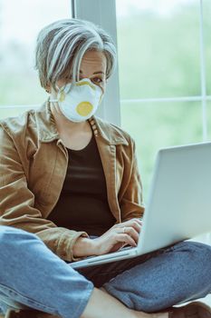 Gray-haired woman in mask fp1 looks at camera using computer while sitting tailor-fashion with crossed legs on a windowsill. Quarantine concept. Tinted image.