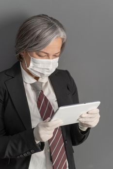 Gray-haired businesswoman in protective mask and gloves using digital tablet. Aged lady holds tablet computer looking at screen. Close up portrait.