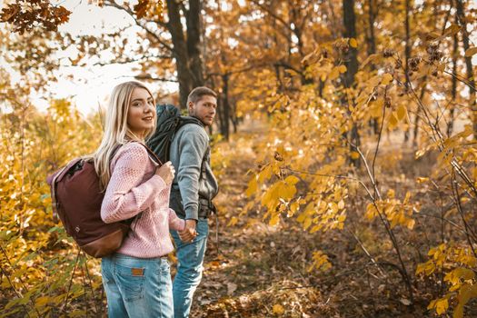 Smiling man and woman in love with backpacks looked back at the camera, happy couple of tourists walking along a forest path holding hands, beauty of autumn nature concept.