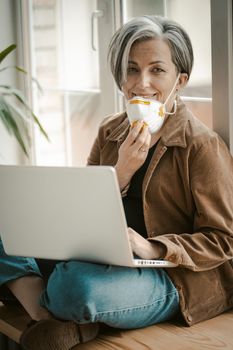 Grey haired woman removes mask affably looking at camera. Caucasian lady uses computer sitting tailor-fashion with crossed legs leaning back against window. Quarantine concept. Tinted image.