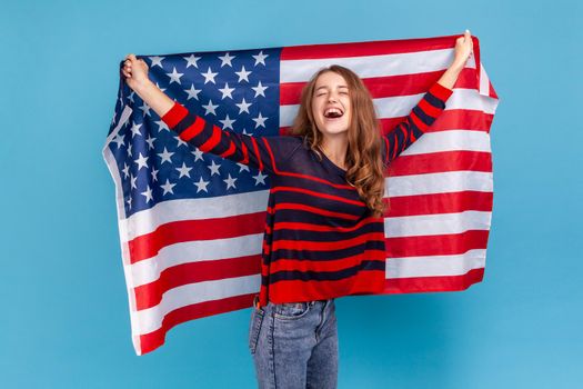 Excited young adult woman wearing striped casual style sweater, holding huge american flag and yelling happily, celebrating national holiday. Indoor studio shot isolated on blue background.
