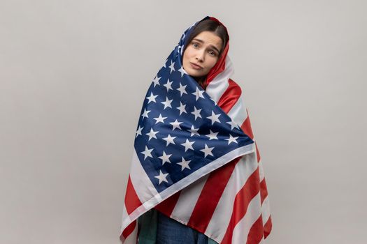 Portrait of relaxed young adult woman standing wrapped in USA flag, celebrating national holiday, looking at camera with pout lips. Indoor studio shot isolated on gray background.