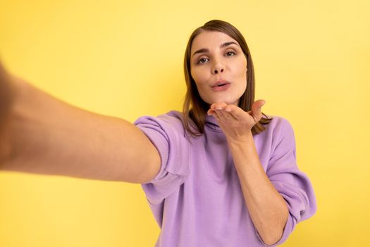 Portrait of romantic young adult woman making point of view photo, sending air kissing, POV, expressing love, wearing purple hoodie. Indoor studio shot isolated on yellow background.