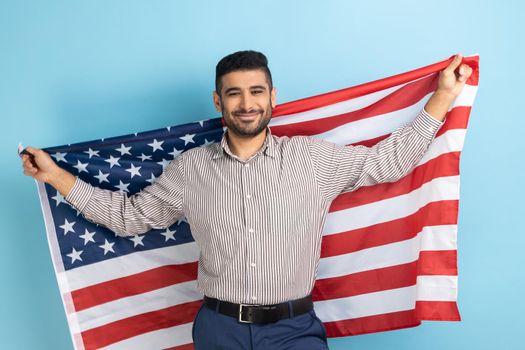 Portrait of smiling businessman holding USA flag and looking at camera with smile, celebrating national holiday, wearing striped shirt. Indoor studio shot isolated on blue background.