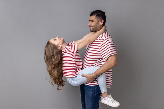 Portrait of delighted satisfied father and daughter in striped T-shirts spending time together, enjoying weekend, dad holding child. Indoor studio shot isolated on gray background.