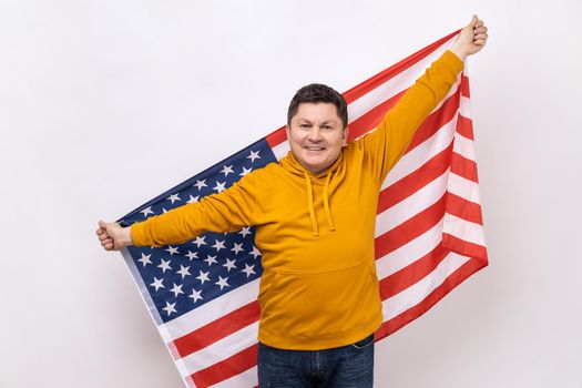 Happy positive middle aged man standing with raised arms, holding USA flag, celebrating national holiday, wearing urban style hoodie. Indoor studio shot isolated on white background.