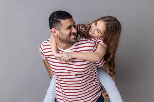 Joyful father giving piggyback ride for his charming daughter, smiling girl looking at her daddy with love, having fun and playing together. Indoor studio shot isolated on gray background.