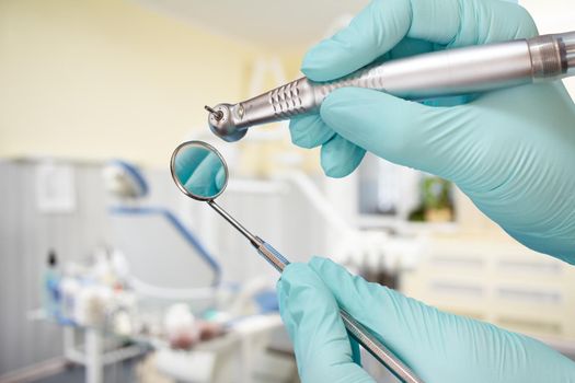 Dentist's hands in pink latex gloves with a high-speed dental handpiece and a mouth mirror with a dental clinic on the background. Medical tools concept.