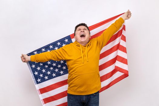 Portrait of middle aged man holding USA flag and screaming happily, looking up, celebrating national holiday, wearing urban style hoodie. Indoor studio shot isolated on white background.