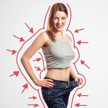 portrait of happy beautiful slim young woman in big jeans and gray top showing successful weight loss, indoor, studio shot, isolated on light gray background, diet concept.