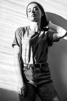 Beautiful woman wearing T-shirt and jeans standing with dreamy romantic expression, keeps eyes closed. Black and white photography, indoor studio shot illuminated by sunlight from window