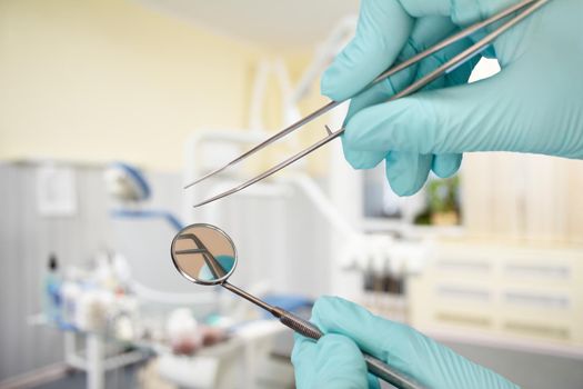 Dentist's hands in latex gloves holding a tweezers and a mouth mirror with a dental clinic on the background. Medical tools concept.