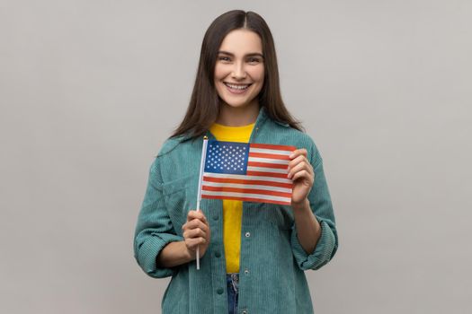 Portrait of young happy satisfied woman standing and holding american flag, celebrating national holiday, wearing casual style jacket. Indoor studio shot isolated on gray background.