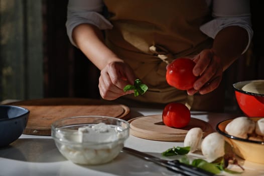 Close-up of housewife's hands. Chef holding ripe tomato and basil leaves while preparing meal in a rustic kitchen. Raw ingredients on chopping board and fresh feta cheese on glass bow on white table