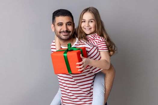 Portrait of happy father and daughter in striped T-shirts standing with present box, kid preparing gift for father's day, expressing positive emotions. Indoor studio shot isolated on gray background.