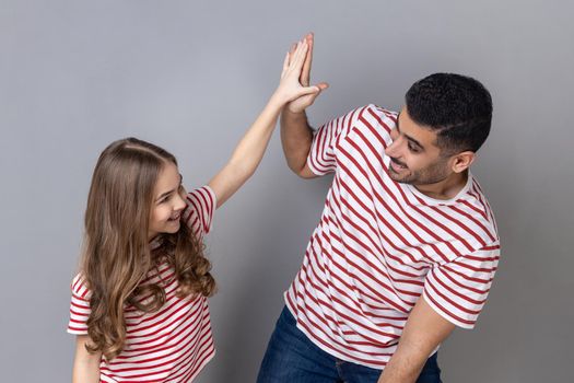 Portrait of playful cheerful father and daughter in striped T-shirts standing together, little girl giving high five to happy dad. Indoor studio shot isolated on gray background.