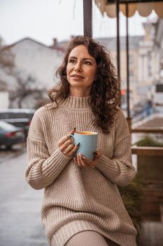 A middle-aged woman in a beige sweater with a blue mug in her hands is in a street cafe on the veranda.