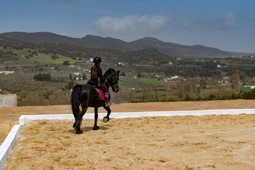 unrecognizable woman riding on a black horse with a mountainous landscape in the background and blue sky